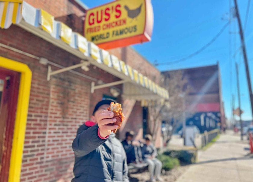 Cannon holding a piece of Gus's chicken in front of the Gus's Chicken restaurant. 