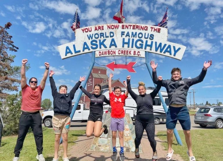 Family jumping in front of the Alaska Highway sign with their RV in the background